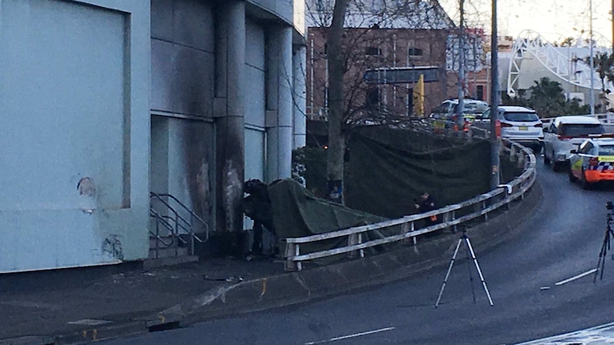 A piece of dark material is draped over the wreckage of a car between a road ramp and a building.