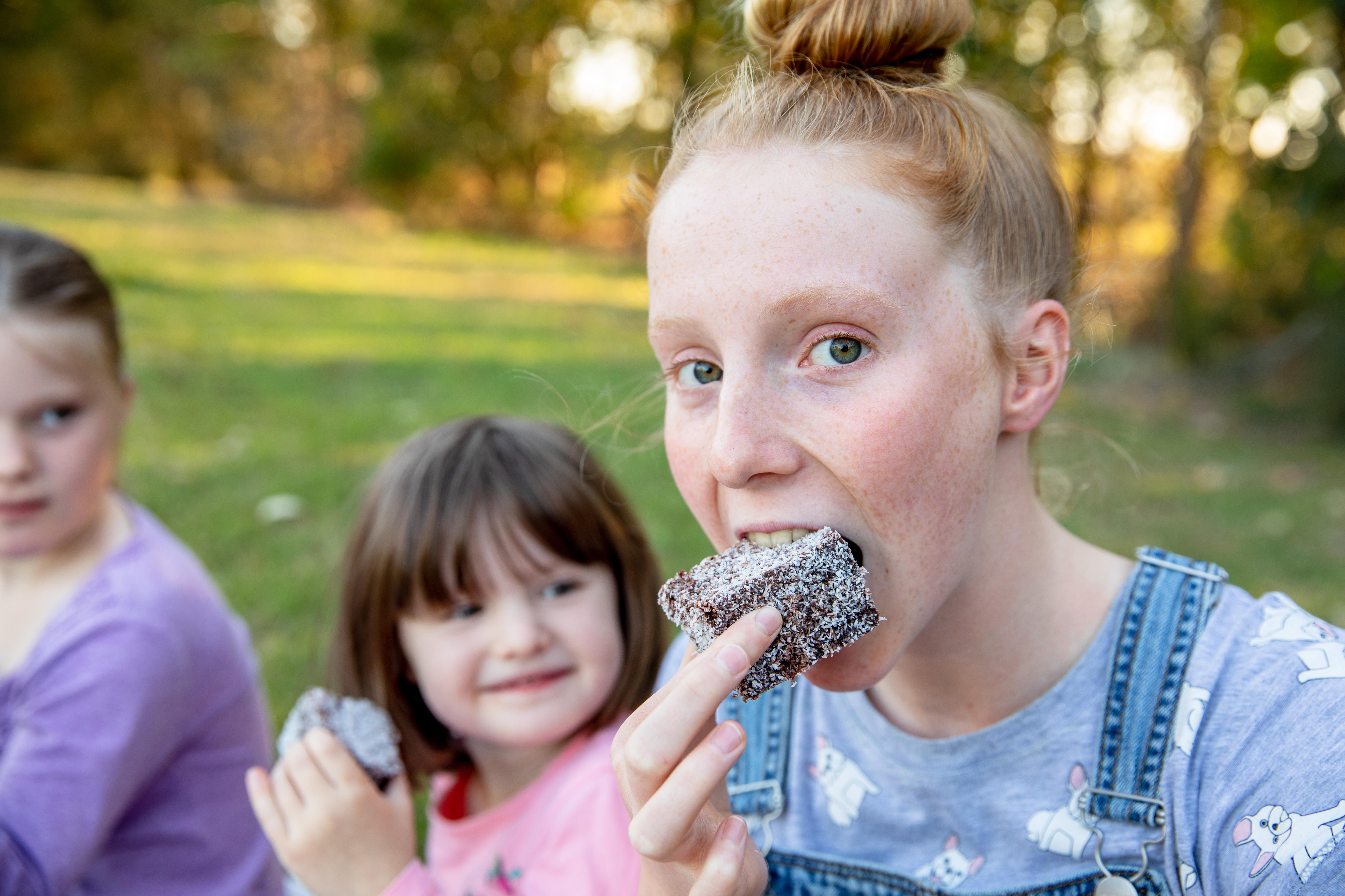 A young girl with red hair looks into the camera as she takes a bite from a square lamington. Two blurred children are behind.