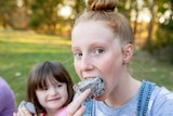 A young girl with red hair looks into the camera as she takes a bite from a square lamington. Two blurred children are behind.