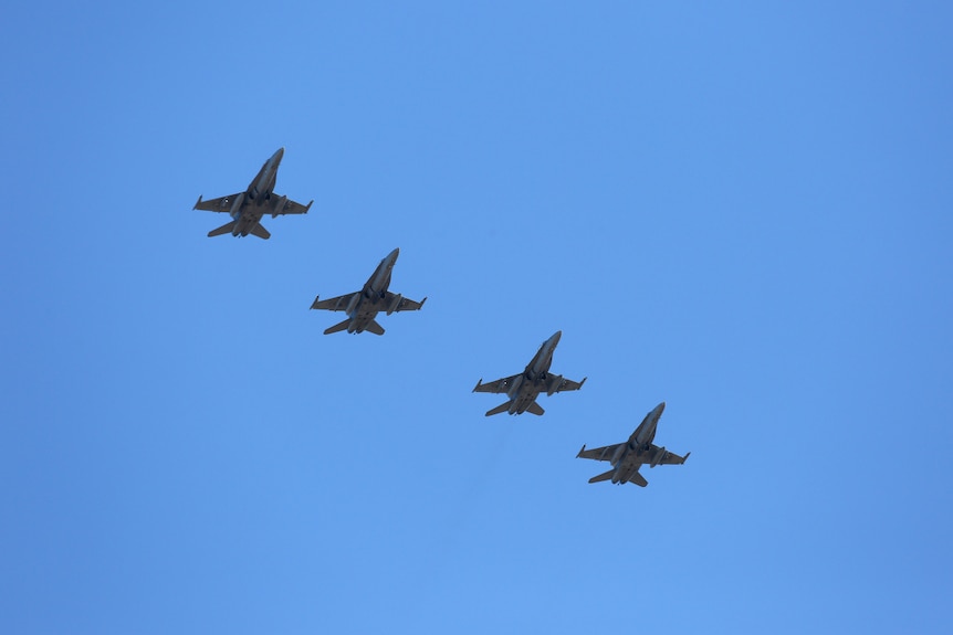 Seen from underneath, four fighter jets fly in a formation across a cloudless blue sky.