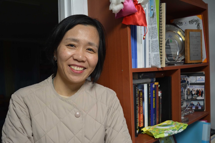 A South Korean woman with bobbed black hair and a beige coat leans against a bookcase and smiles.