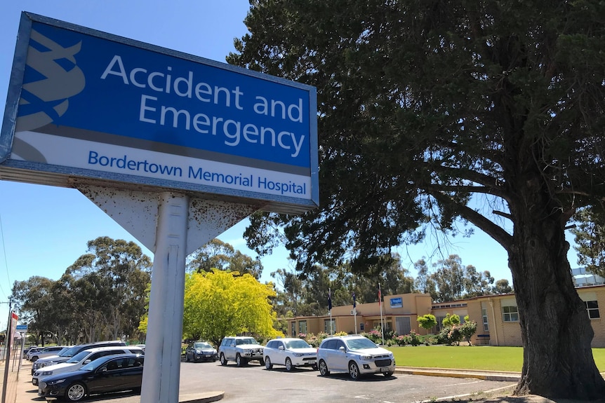 An emergency centre sign with hospital buildings and a tree
