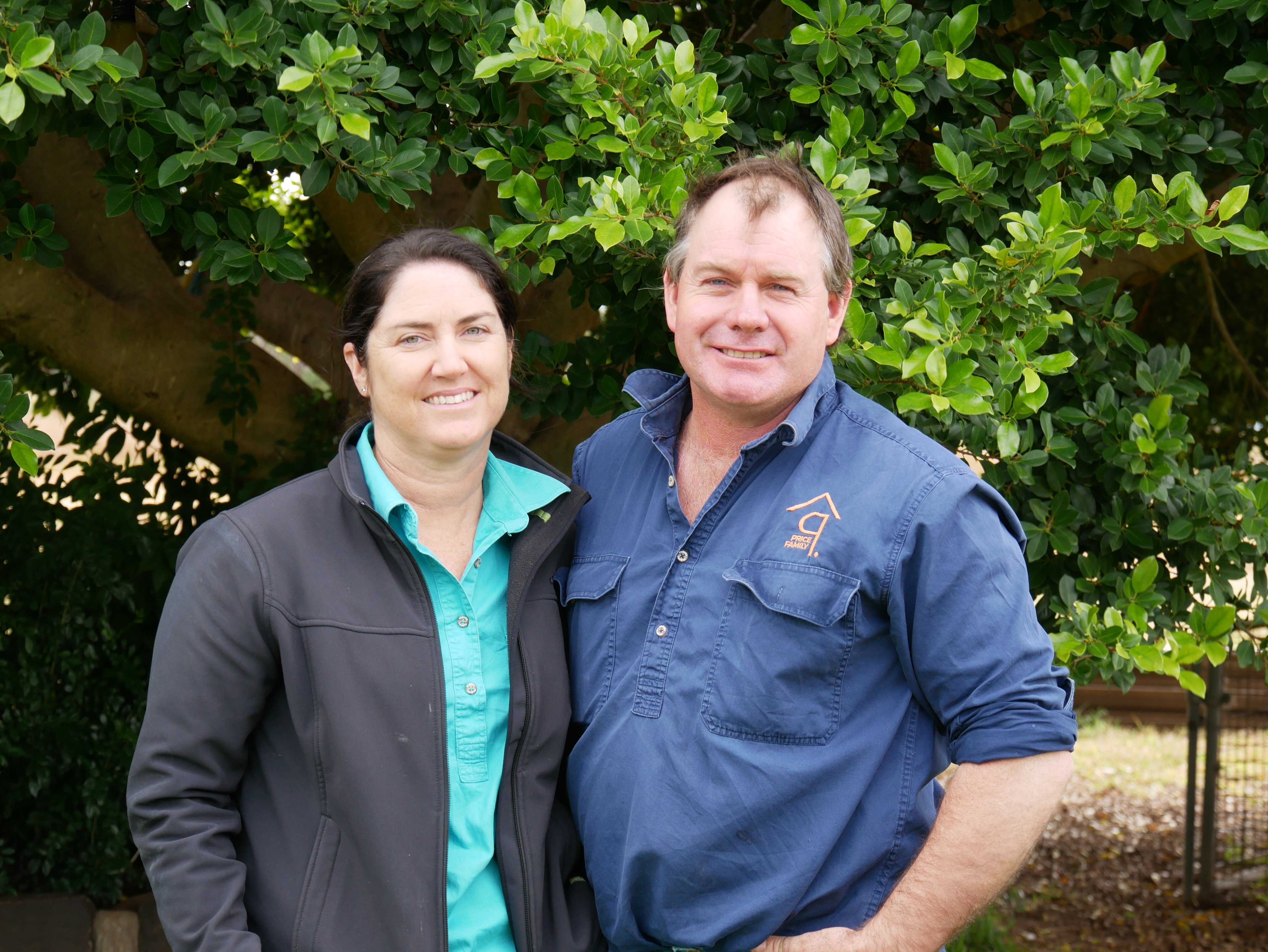 A women and man both in blue workshirts stand next to eachother in front of tree with green leaves