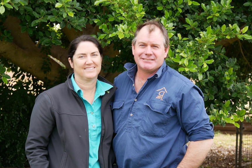 A women and man both in blue workshirts stand next to eachother in front of tree with green leaves