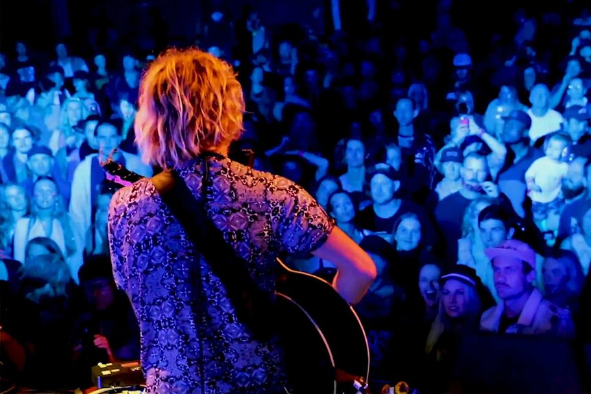 A sea of faces watching a single male viewed from behind playing a guitar