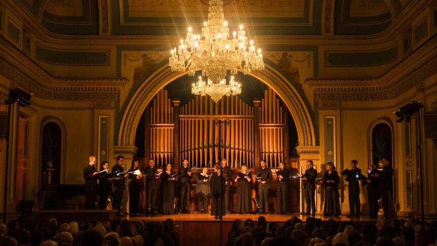 A moodily lit hall with singers dressed in black in performance under a chandelier.