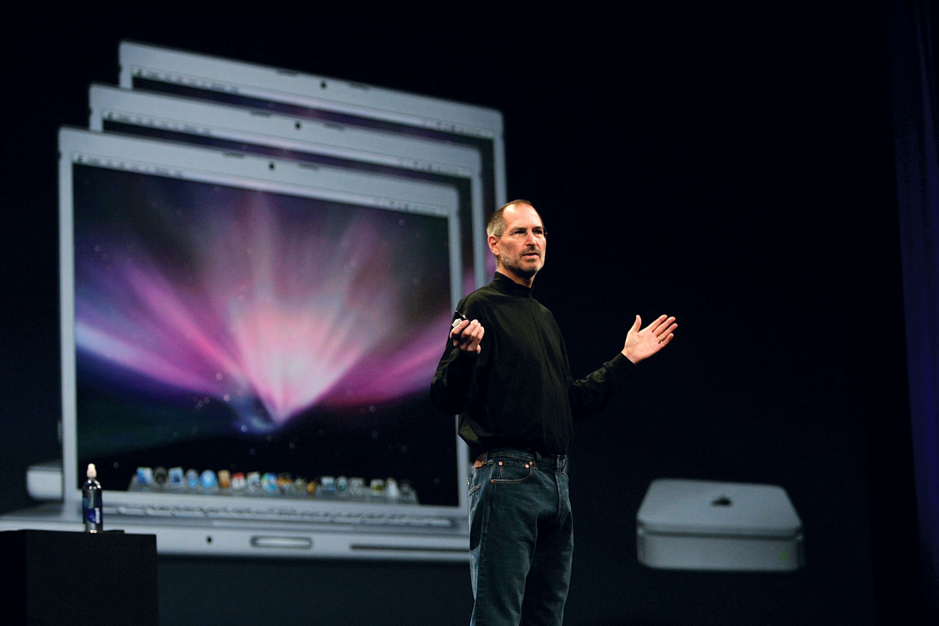 A man wearing a black turtleneck top stands on stage with his hands outstretched, a computer displayed on a screen behind him.