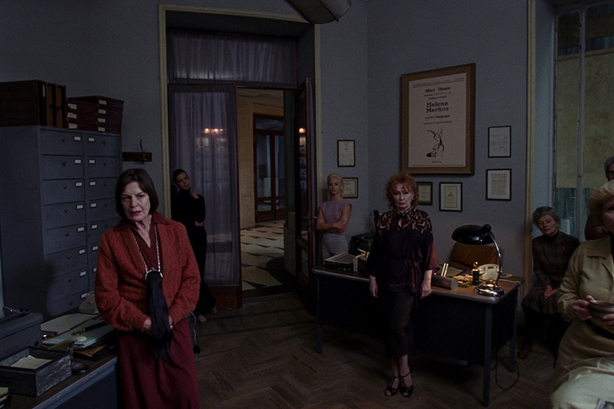 Colour still of a seven women standing in various spots in a dimly lit office space in 2018 film Suspiria.