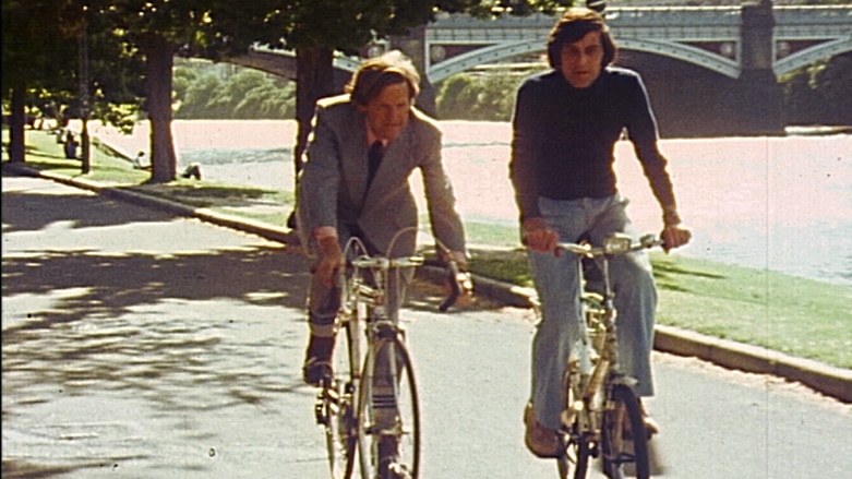 Two men in 1970s style clothing ride bicycles along a riverside path with bridge in background.
