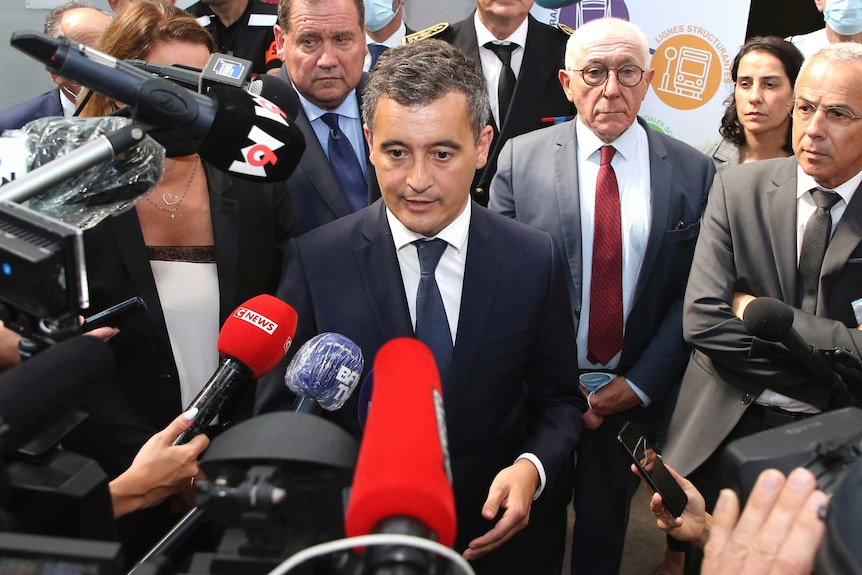 A man wearing a suit speaks in front of a media crowd.