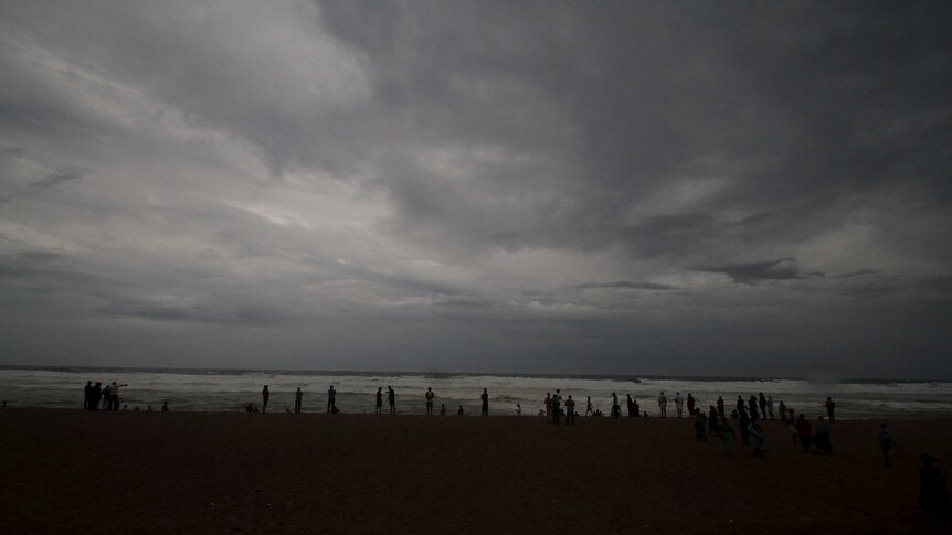About 20 people stand silhouetted in the far distance on a beach with big looming grey clouds above them.