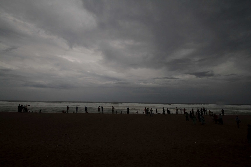 About 20 people stand silhouetted in the far distance on a beach with big looming grey clouds above them.