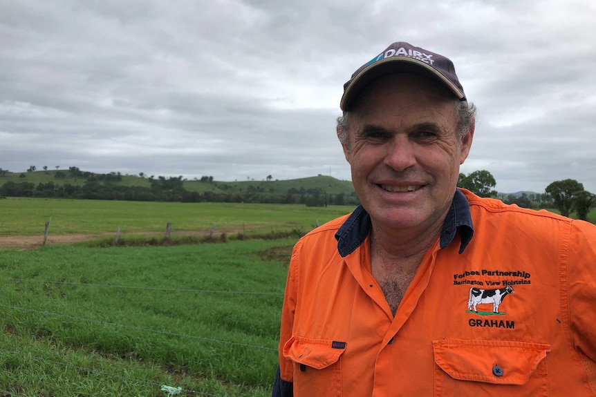 A farmer in an orange shirt and black cap stands in a lushly green paddock, smiling at the camera