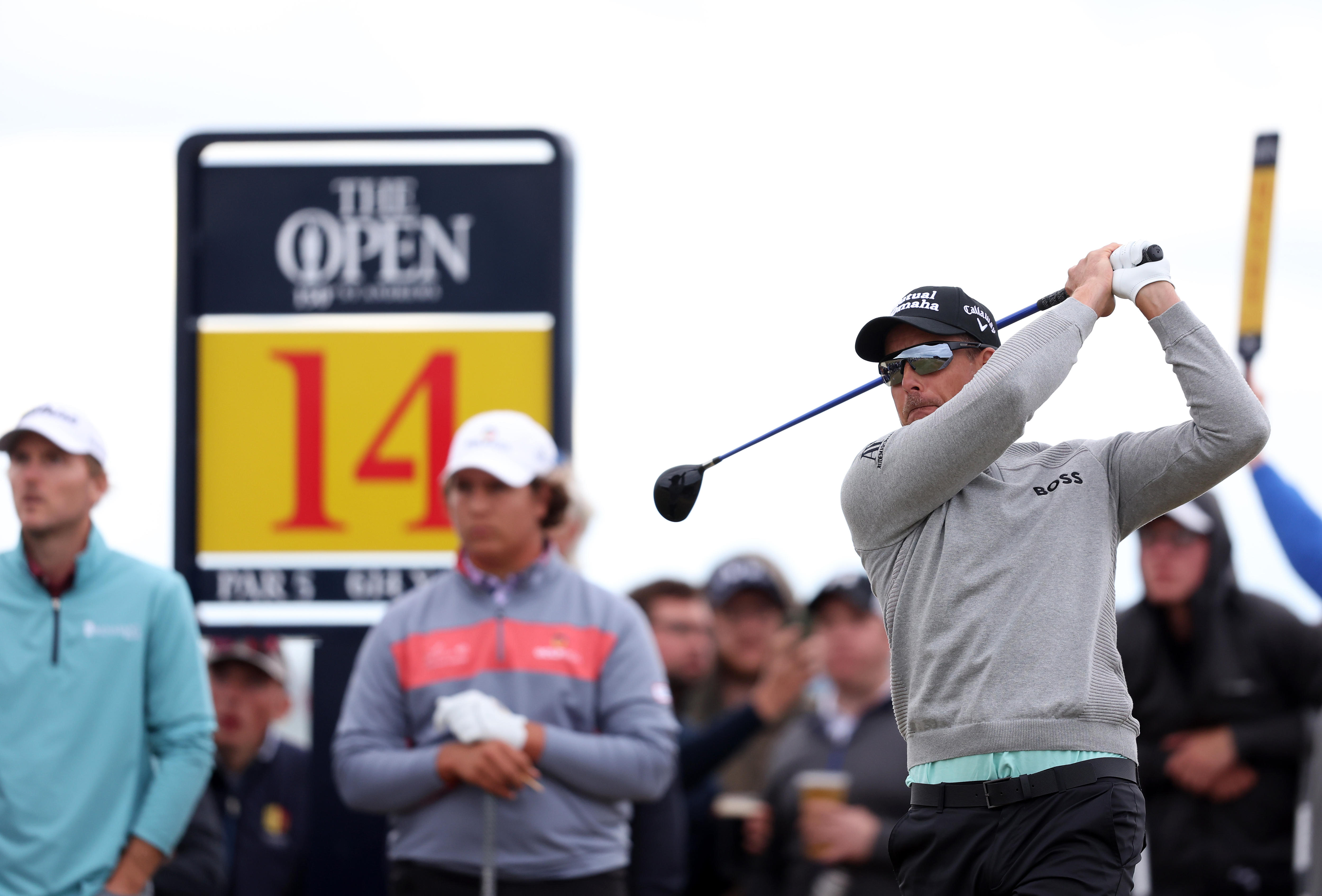 A sweater-wearing golfer hits off the tee with a sign saying "The Open, 14" in the background. 