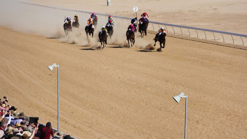 Horses racing along a dusty track with onlookers watching on.