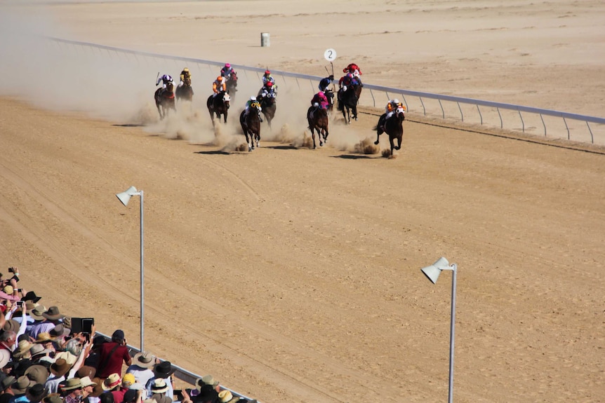 Horses racing along a dusty track with onlookers watching on.