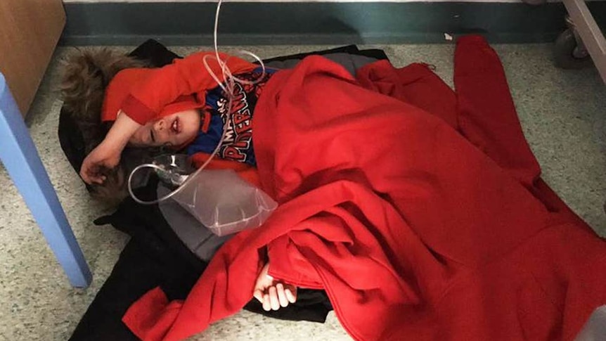 A small child sleeps on the floor of a UK hospital wrapped in coats and jumpers with medical tubes around him