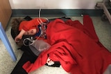 A small child sleeps on the floor of a UK hospital wrapped in coats and jumpers with medical tubes around him