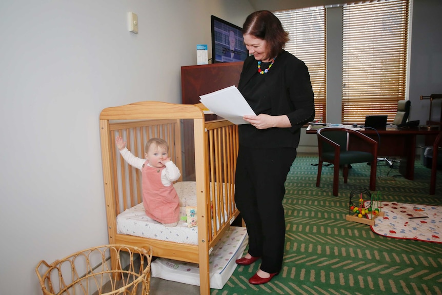 Lisa Chesters smiles wile standing next to her cot, which her daughter is in