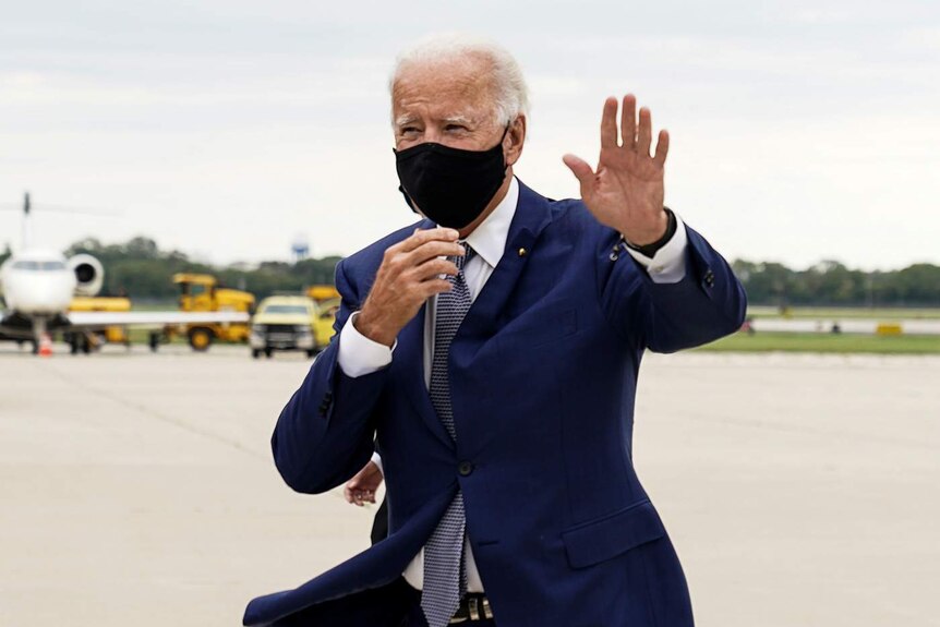 You view Joe Biden in a navy suit with a black face mask walking on an airport runway on an overcast day.