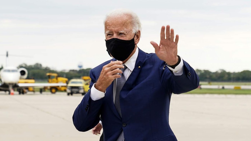 You view Joe Biden in a navy suit with a black face mask walking on an airport runway on an overcast day.