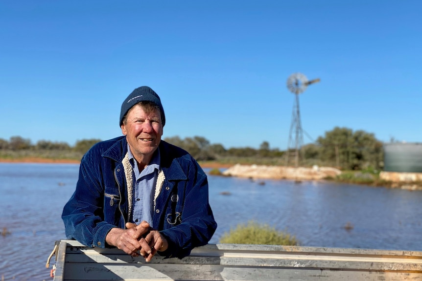 A man leans over the try of a ute. He is wearing a beanie and a black jumper. Behind him is a pool of water and a windmill. 