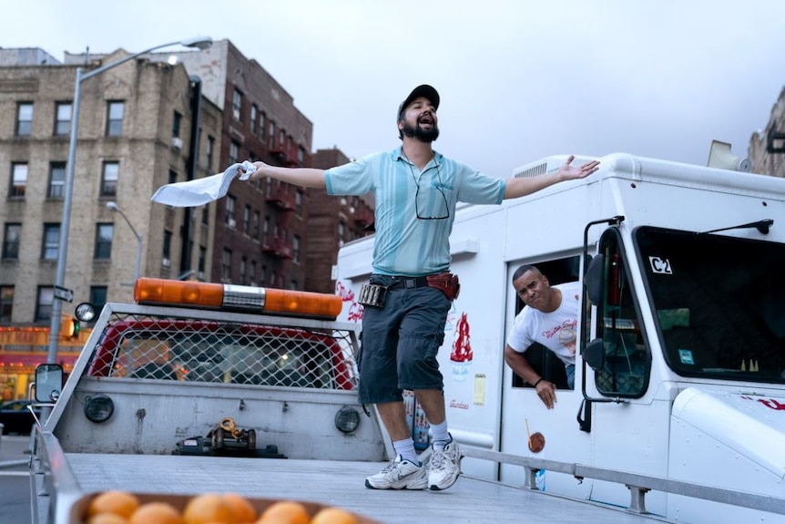 A man sings and dance on top of a truck carry tray while an ice cream driver looks on annoyed.