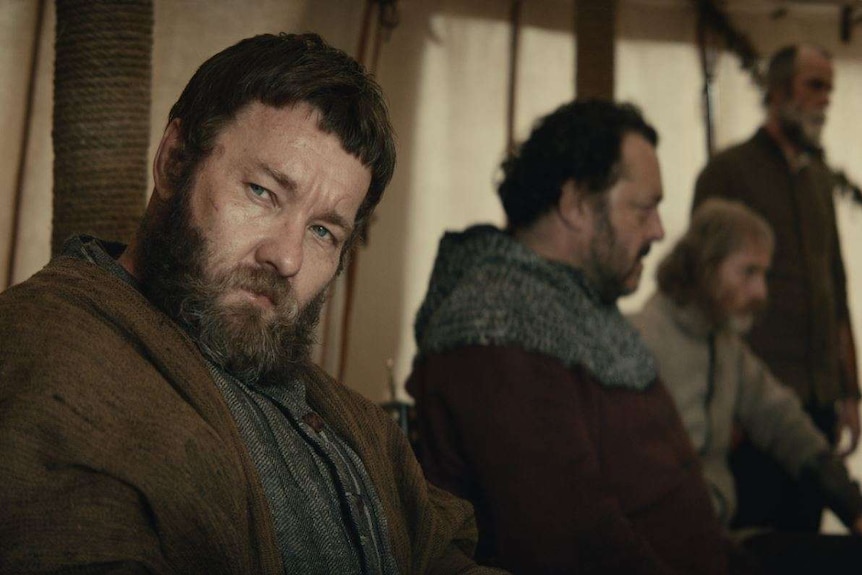 The actor Joel Edgerton in medieval garb and big beard, sitting in a tent with men dressed similarly in the background