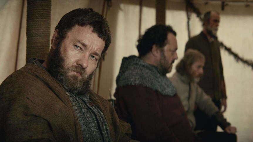 The actor Joel Edgerton in medieval garb and big beard, sitting in a tent with men dressed similarly in the background