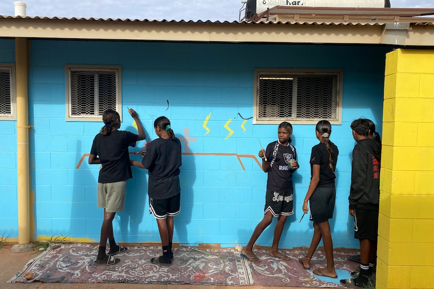 Five aboriginal girls stand in front of blue wall they are painting.