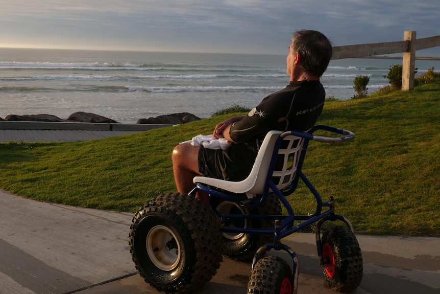 Man draped in morning sun sits in rashie in wheelchair looking at ocean