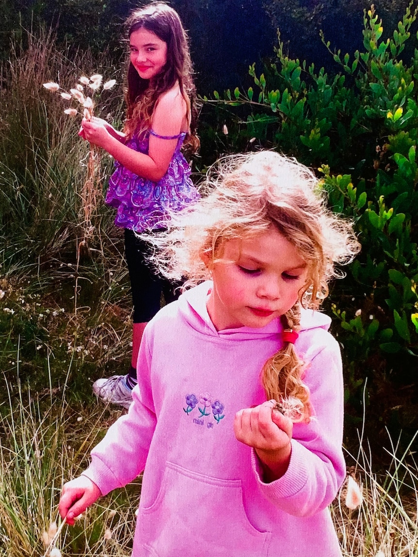 Smiling young girl in purple singlet, and a younger girl in pink jumper stand in long grass and trees. Young girl looks at hand.
