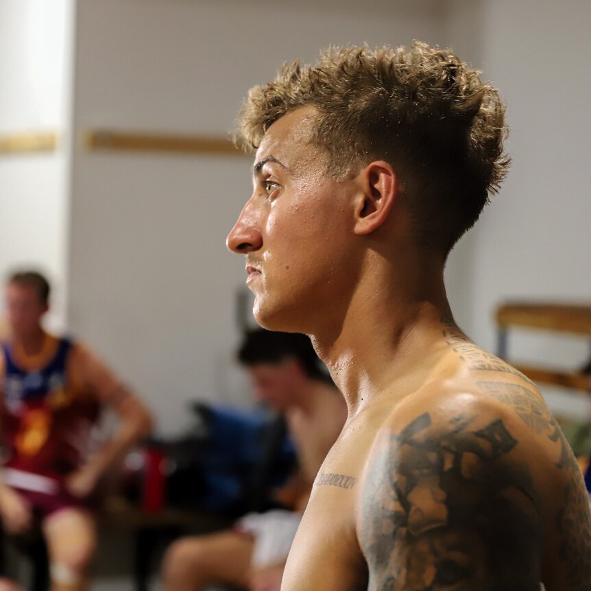 Shirtless young man stares intently inside football clubrooms with other players in background