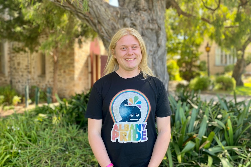 A young non-binary person with long blonde hair, wearing a T-shirt with a cartoon whale on it, stands in front of a tree
