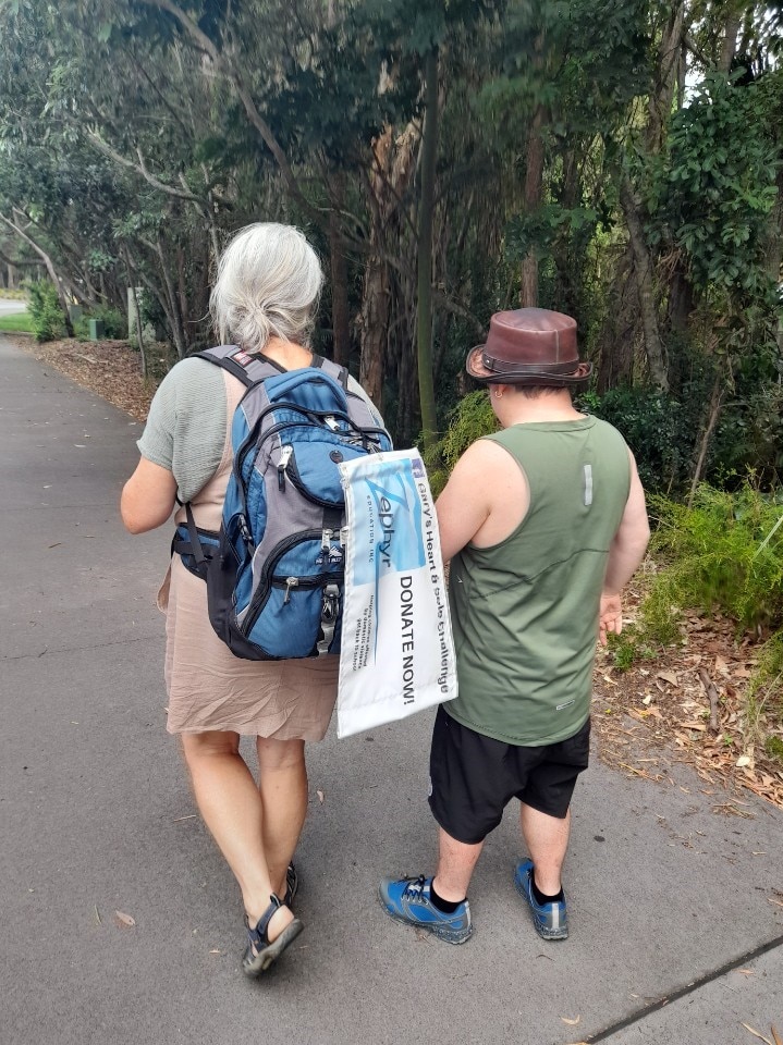 The backs of a mother and her adult son walking on a concrete path with trees in the background.