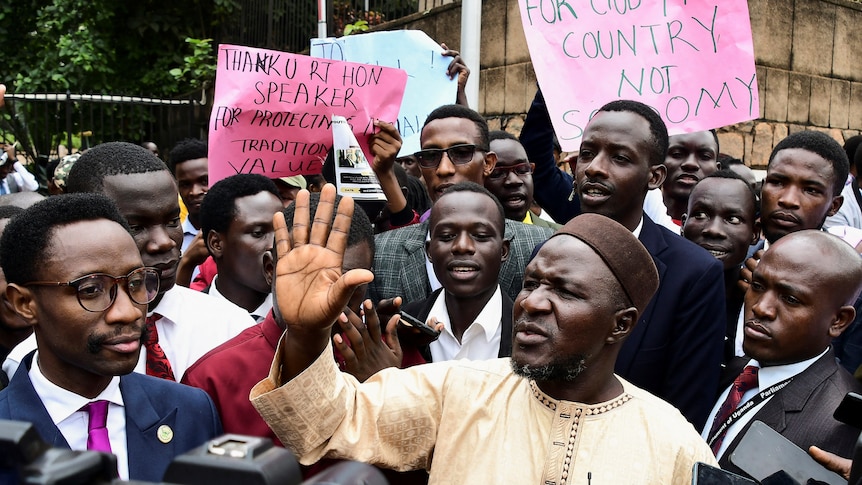 A group of young African men, many wearing suits and holding signs, stand in a group and speak to journalists.