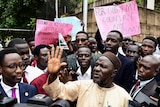 A group of young African men, many wearing suits and holding signs, stand in a group and speak to journalists.
