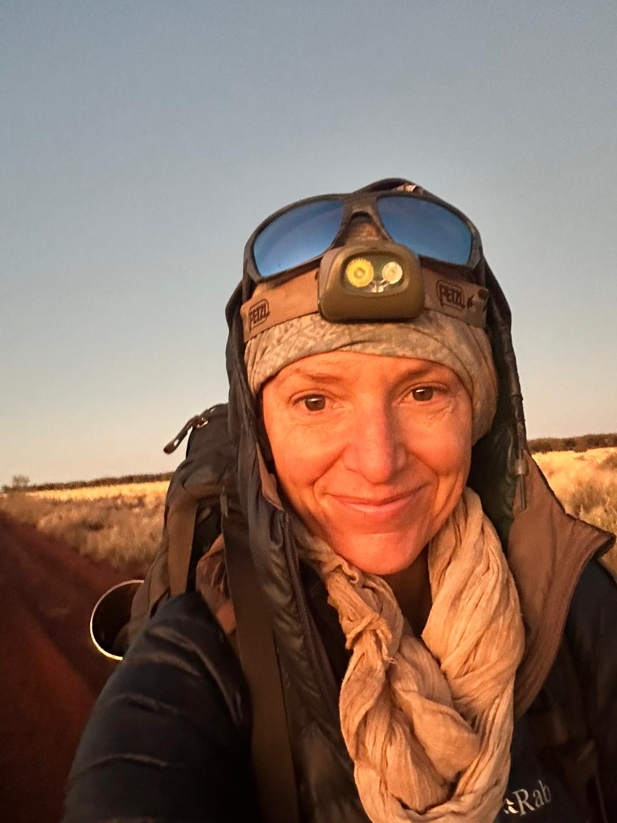 A smiling woman in adventure gear, wears goggles on her head, a beanie, an equipment strapped on, carrying backpack.