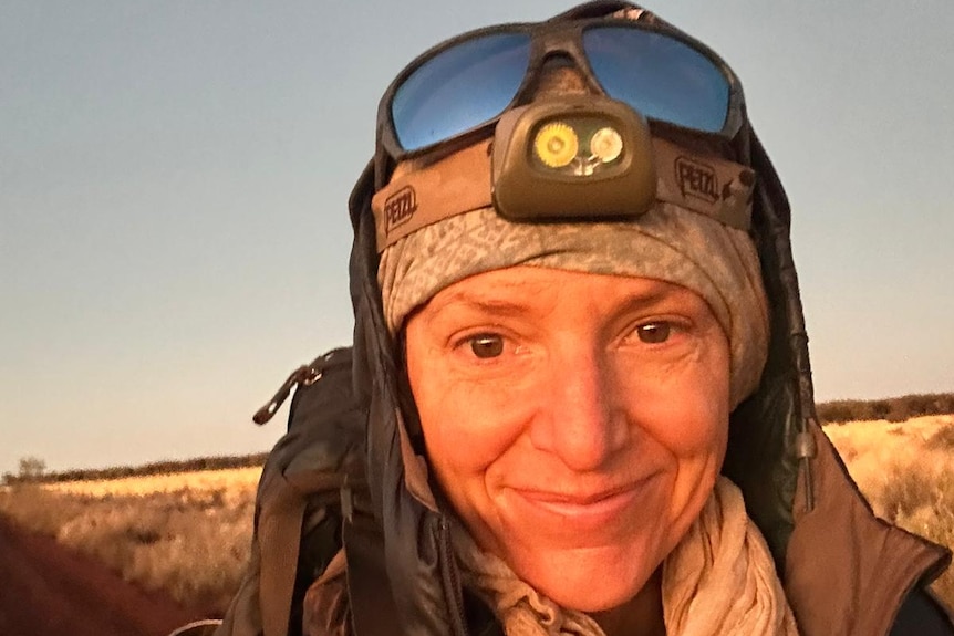 A smiling woman in adventure gear, wears goggles on her head, a beanie, an equipment strapped on, carrying backpack.