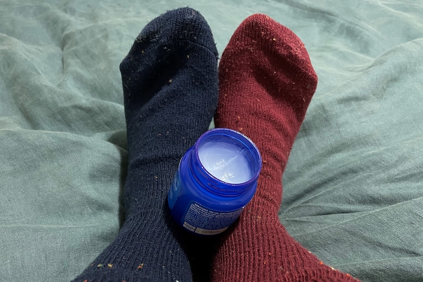 An open jar of vapour rub nestled in the feet of someone wearing a navy blue and a maroon sock. 