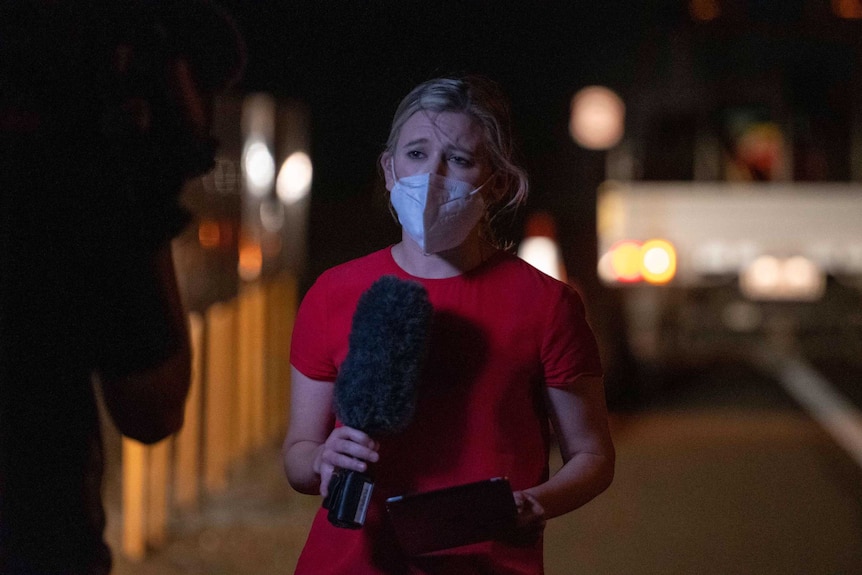 Woman holding microphone wearing mask and standing in front of camera at night with blurred lights in background.