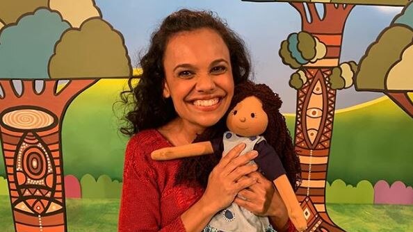 Miranda Tapsell smiles as she clutches Kiya with a forest mural behind her. She wears a red jumper and Kiya is in a dress.