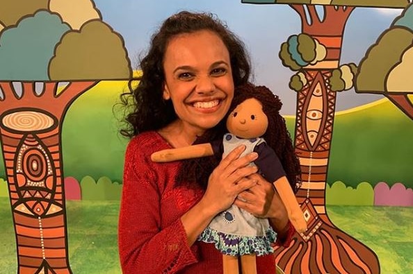 Miranda Tapsell smiles as she clutches Kiya with a forest mural behind her. She wears a red jumper and Kiya is in a dress.