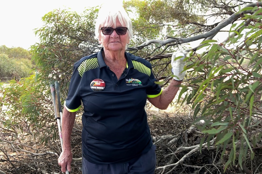 Woman in navy shirt wearing white golf glove holding up golf ball in hand and holding golf srick in other in front of scrub tree