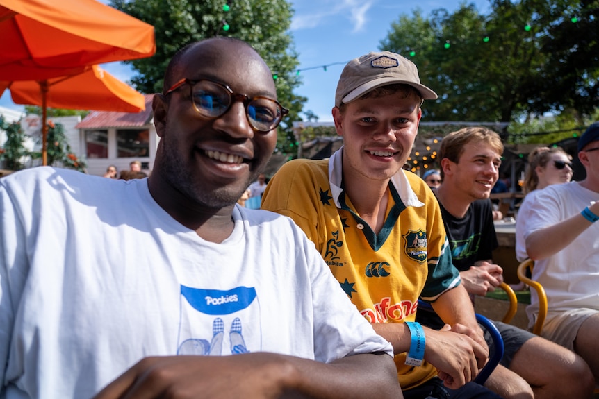 Two smiling young men sitting at the back of a beer garden in London.  One is wearing a Wallabies jersey.
