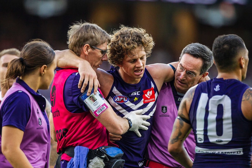 Nat Fyfe with his arms around the shoulders of two Dockers support staff, as he is helped from the field while injured.
