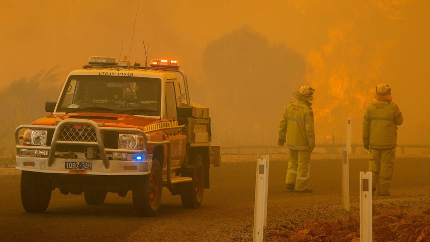 Two firefighters near a four-wheel drive emergency services vehicle look on as a bushfire rages near a road.