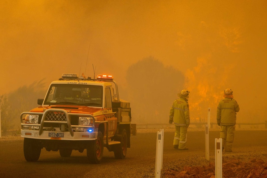 Two firefighters near a four-wheel drive emergency services vehicle look on as a bushfire rages near a road.