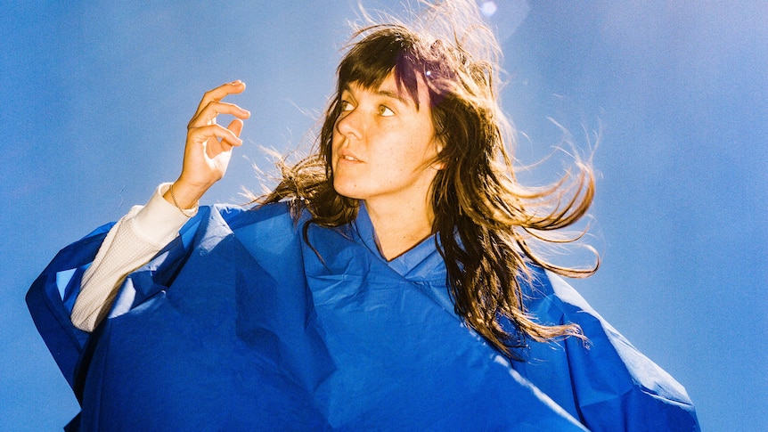 Courtney Barnett stands in a blue raincoat lifting her right hand against a clear blue sky with lens flare