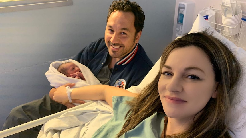 A woman with dark hair lies on a hospital bed next to a man with dark hair who holds a baby wrapped in a blanket.